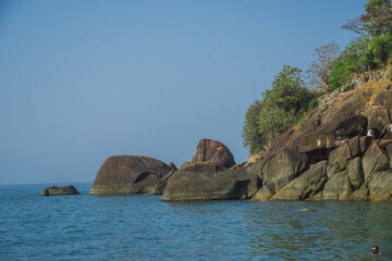 Man wearing sun glass enjoying and swimming into the sea near shore. Big rocks and green forest background on an island. Butterfly beach in Goa, India. Blue ocean water with huge rocks and nearby hill