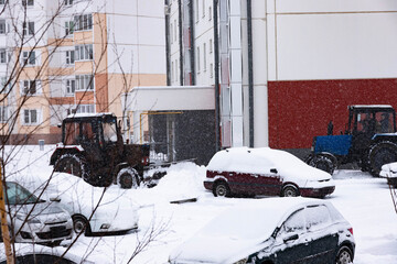 The tractor clears the road from snow in winter during a snowfall. There is a strong blizzard
