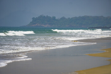 Beautiful sandy beach in Goa, India. Low tides in Sea on a sunny day. Green forest hills near sea shore. Brown sandy beach and white waves in water. Sun reflection in sand when water hits the sand.