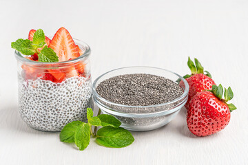 Chia seeds in glass bowl used as ingredient for healthy natural pudding made with vegetarian plant based milk decorated with sliced juicy strawberries topping and mint leaf served on white table