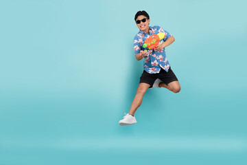 Fototapeta na wymiar Handsome Asian young man in summer clothes is jumping and using a water gun on a blue background. Songkran Festival in Thailand