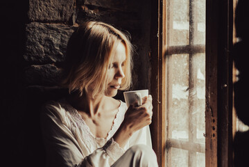 Obraz premium A woman with a pensive look drinks coffee at the window. Middle age woman in a white shirt. Sad look, thoughts.