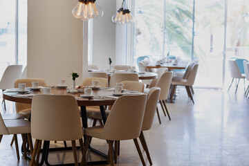 Modern dining area with multiple tables set with white dishes and silverware. Each table is surrounded by beige chairs. Pendant lights hang from the ceiling. Large windows reveal lush greenery outside