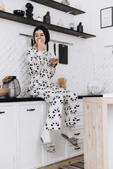 Fototapeta na wymiar Cheerful woman standing in bright kitchen smiling eat candy, dressed in patterned pajama set. The kitchen counter arranged with ingredients, utensils 