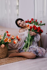 A woman poses near bed, surrounded by vibrant fresh bouquets bunch of red tulips placed around the room, domestic atmosphere. Spring International Women's Day, 8th of March. Holiday, festive 
