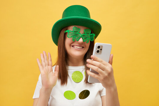St. Patrick's Day celebration full of fun and joy. Smiling woman wearing festive green hat and shamrock glasses standing isolated over yellow background having video call waving hello gesture