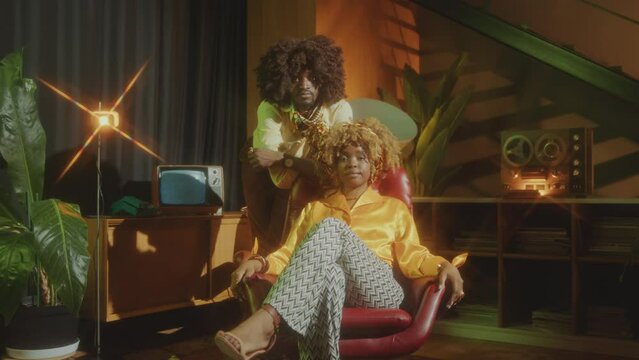 Young Attractive African American Woman Sitting In Chair As Boyfriend Standing Behind, Both Posing At Camera With Confidence In Old-fashioned Studio. Video Portrait, Retro Glow Effect