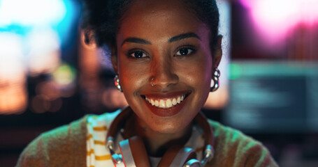 Portrait of a Stylish Young Black Woman with Headphones, Looking at Camera, and Smiling in a Creative Office. Beautiful Diverse Female Working for her Dream Agency, Feeling Empowered