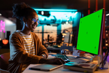 Portrait of Creative Black Woman Sitting at Her Desk Using Desktop Computer with Mock-up Green Screen. Female Specialist Working at Night in Game Development Startup, Finishing Details on a Project