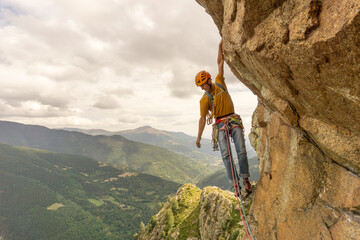 A man is climbing a rock wall with a yellow shirt and an orange helmet. The scene is set in a...