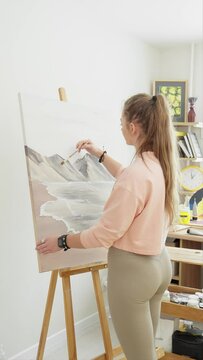 Artist creating artwork in art studio. Modern artwork paint on canvas in studio. Talented female artist indoors. Artist paints with acrylic paint. Landscape, sea, mountains. Vertical footage. 4K, UHD