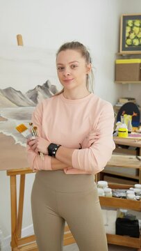 Portrait of talented young female artist looking at camera with artwork at background. Woman crosses arms while holding brushes. Talented woman artist indoors. Vertical footage for stories. 4K, UHD