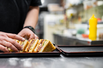 chef prepares fresh sandwiches on a black tray in a professional kitchen. The neatly arranged sandwiches feature multiple layers of ingredients, including meat and vegetables between slices of bread