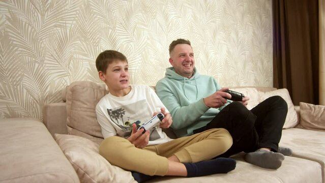 A father spends time with his little son, sitting on the couch and playing a game console. Weekend activities, free time, home entertainment and video games concept.