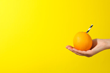 Orange with cocktail straw on hand on yellow background, space for text - obrazy, fototapety, plakaty