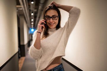 Stylish woman wearing sweater and glasses talking on smartphone in hotel corridor - obrazy, fototapety, plakaty