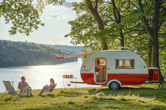 A serene scene of a couple relaxing beside a lake with a vintage red and white caravan surrounded by nature's tranquility