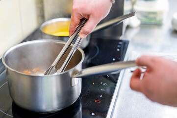 Close-up of hands using tongs to stir food in a stainless steel pot on an induction stove. Another pot with a yellowish liquid is visible in the background