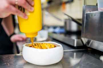 Chef in a professional kitchen drizzling mustard from a yellow bottle onto a dish, with blurred kitchen equipment in the background.