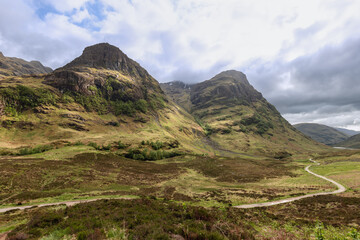 A winding path through the Glen Coe valley invites exploration amidst the Three Sisters, under a dynamic sky that casts a dramatic light over the Scottish highlands - obrazy, fototapety, plakaty