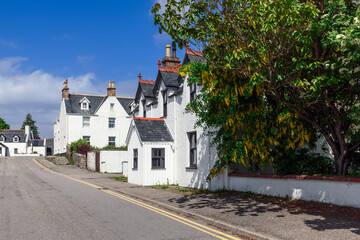 Fototapeta na wymiar Historic white buildings with red trim line a quiet street in Ullapool, framed by lush greenery and blooming yellow laburnum under a blue sky. Highland Council, Scotland
