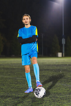 young Caucasian girl in a blue sportswear posing with a soccer ball in a stadium, full vertical shot. High quality photo