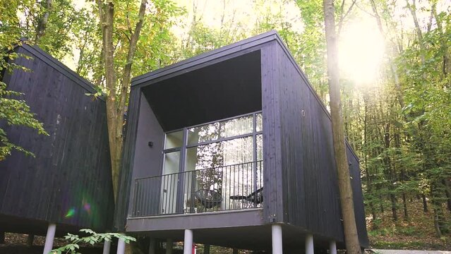 Exterior view of a small vacation or recreation house in dark colors with large windows and a balcony or terrace between trees in the forest