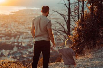 A back shot of a father holding his son's hand, gazing into the distance atop a hill overlooking the city.