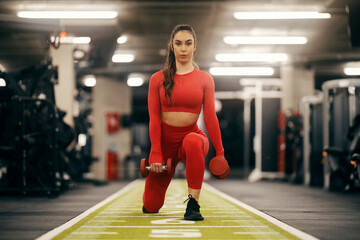 A fit sportswoman is doing lunges with dumbbells in her hands in a gym.