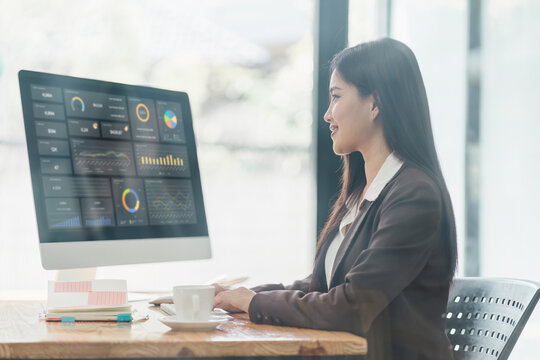 Asian businesswoman or accountant are analyzing data charts, graphs, and a dashboard on desktop screen in order to prepare a statistical report and discuss financial data in an office.
