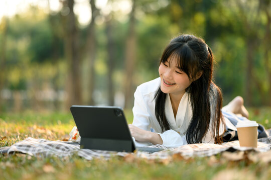 Portrait of pretty young Asian woman lying on picnic blanker and using digital tablet
