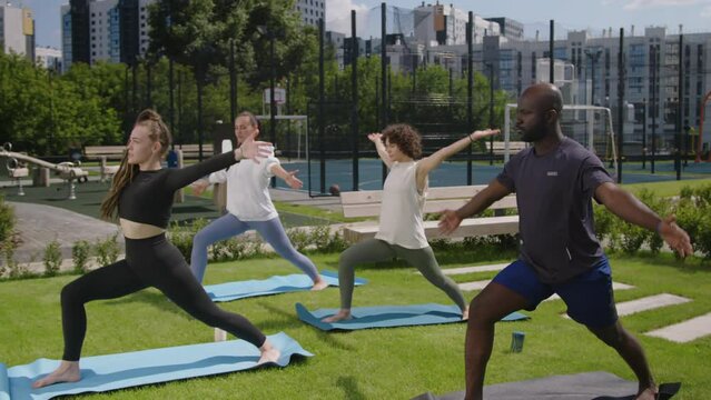Side full footage of diverse people repeating asanas after female yogi during outdoor yoga workout in rooftop park, doing warrior pose or virabhadrasana