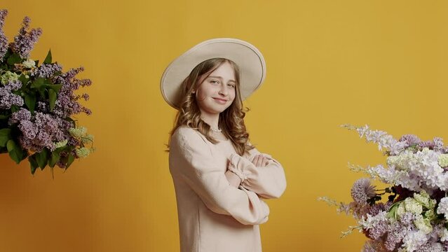 Beauty teenager girl 13 years in a beige dress and hat is happy and smiling and looking at camera on a yellow background with natural flowers in the studio. Cheerful teenage girl looking at camera.