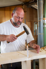 Tischler arbeitet in seiner Werkstatt, schlägt einen Holzdübel ein mit Leim - 756349076