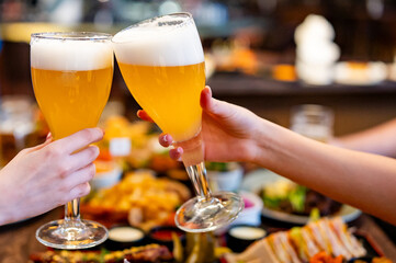 Two hands clinking beer glasses in a cozy indoor setting. Assorted food items are visible on a wooden table in the blurred background