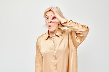 Surprised woman in beige blouse making OK gesture over eye, white background.