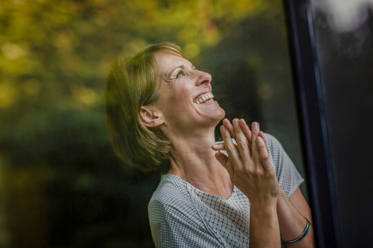 40 j&auml;hrige gl&uuml;ckliche Frau am Fenster Blick nach draussen, Spiegelungen Laubbaum, Bayern, Deutschland