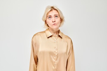 Confused young woman in beige blouse standing against light grey background with puzzled expression.