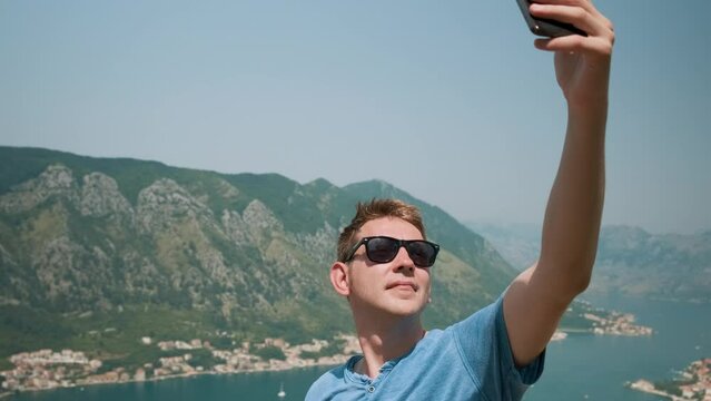 Man tourist in sunglasses take a selfie with Kotor bay, Montenegro on background standing on Lovcen mountain. Handsome male traveler on summer vacation taking selfies with landmark