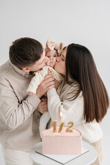 Obraz na płótnie Canvas Dad and Mom kiss cute baby isolated on white wall. Father, mother hold hands funny little kid. Creative birthday cake with candle. Family celebrates six month old baby girl birthday party closeup.