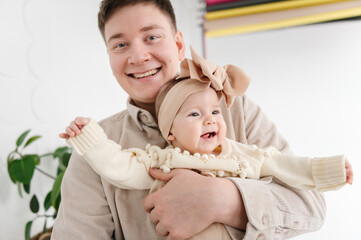 Dad holds in hands a 6 month old little girl. Father hugging his daughter. Cute baby in dad's arms closeup. Portrait of daddy with an adorable baby girl with bow in hair at home.