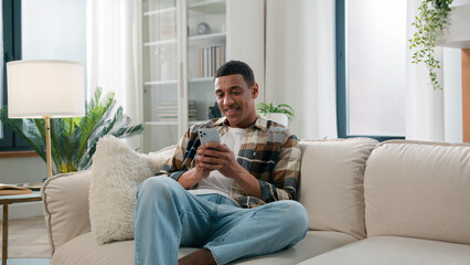 Back view African American guy enter living room walk at home approach go to sofa sit on couch browsing mobile phone man scrolling smartphone social media indoors using cellphone app chatting relaxing