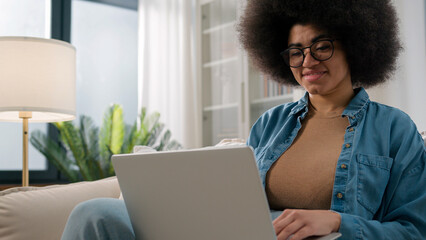 Busy African American woman using pc at home freelancer female user on couch sofa working with...