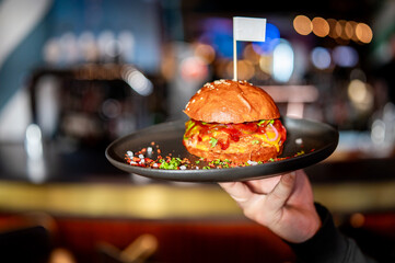 Close-up of a person holding a juicy burger with a flag toothpick, served on a black plate in a vibrant bar setting. The burger has visible layers of lettuce, cheese, and sauce