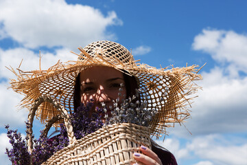 Portrait of a young beautiful woman with a bouquet of lavender in a straw hat against the background of a sunny cloudy sky. Woman enjoys the scent of freshly cut flowers in a wicker basket.