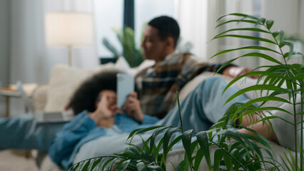 View through house plant African American happy family couple relaxing on couch man working playing...