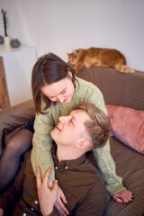 a young couple is hugging on the sofa in the living room, a red cat is sleeping on the back of the...