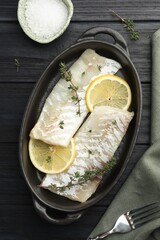 Fresh raw cod fillets with thyme and lemon in baking dish on black wooden table, flat lay