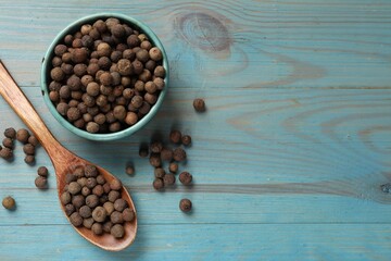 Aromatic allspice pepper grains in bowl and spoon on light blue wooden table, flat lay. Space for...