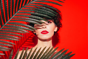 Fashion beauty portrait of young confident brunette woman with evening stylish makeup and perfect clean skin. Sexy model with curly hair posing in studio. With red bright lips. Tropical palm leaf